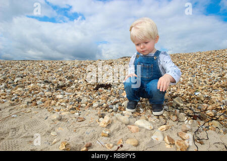 A blonde male toddler in dungarees squatting and looking at stones on a quiet beach Stock Photo