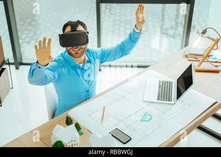 Virtual reality. Emotional progressive engineer putting his hands up and smiling while using virtual reality glasses in his work Stock Photo