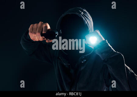 Armed robbery, criminal with hoodie holding gun and torchlight and attacking in dark, low key selective focus Stock Photo