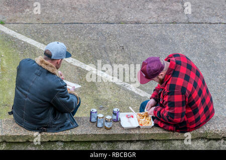 Two men sitting on a low wall outside eating fish and chips and drinking cans of bitter shandy in Wells-next-the-Sea, Norfolk. Stock Photo