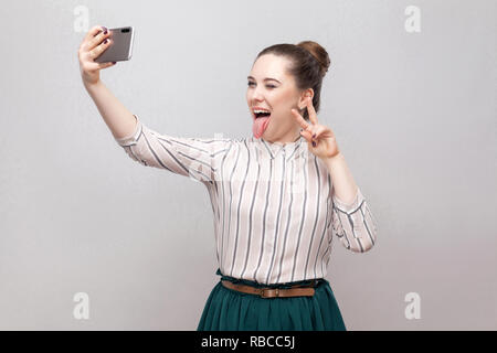 Selfie time! Portrait of happy foolish joyful attractive blogger woman wearing in striped shirt standing, winking and showing tongue and making selfie
