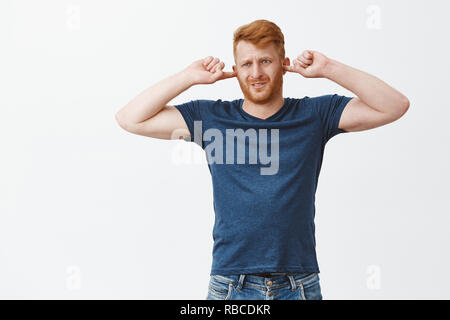 Hate when wife sings in shower. Indoor shot of displeased ginger man feeling discomfort, frowning and grimacing, covering ears with earplugs and gazing aside, hearing unpleasant noise or sound Stock Photo