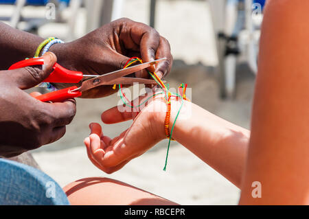 African ethnicity man cutting an extra threads of just braided bracelet from colored threads with scissors on caucasian ethnicity mans wrist on the be Stock Photo