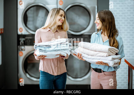 Two young women standing together with clean clothes in the self serviced laundry with dryer machines on the background Stock Photo