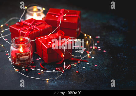 Gift red boxes with red bow ribbons, candles, lights garland and hearts on dark background for Valentines day. Space for text. Stock Photo