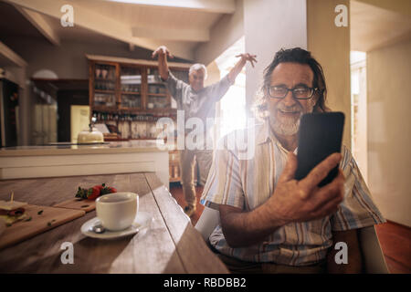Happy senior man taking selfie with friend standing at back making funny gestures.  Two elderly men taking selfie with mobile phone at home. Stock Photo