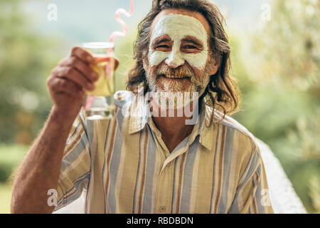 Bearded senior man with clay facial mask holding a glass of juice in hand. Man having spa facial treatment raising his juice glass at camera. Stock Photo