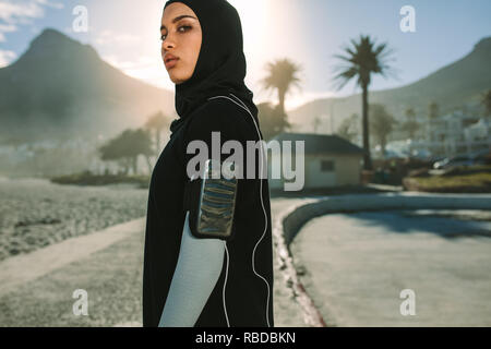 Islamic woman in sportswear standing outdoors in city on sunny day. Muslim female runner in hijab listening to music from mobile phone and looking at Stock Photo