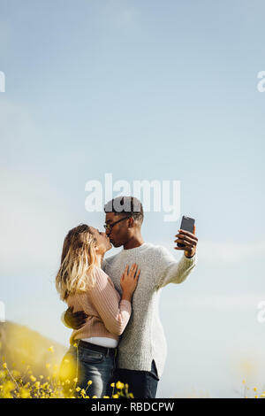 Loving interracial couple kissing and taking self portrait outdoors. Romantic couple taking selfie with their mobile phone. Stock Photo