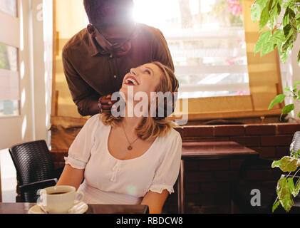 Young man standing behind his girlfriend and laughing in cafe. Couple on date with man surprising his woman at coffee shop. Stock Photo