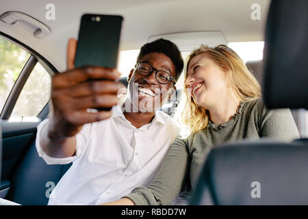 Cheerful interracial couple sitting on back seat of a car and taking selfie with mobile phone. Smiling man with girlfriend taking selfie while traveli Stock Photo