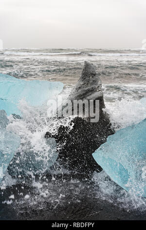 Ice blocks in blue and dark shades on a black beach with strong surf, spray of a wave splashes over the ice, waves roll in the background - Location:  Stock Photo