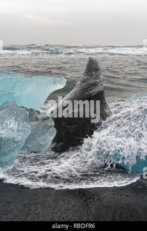 Ice blocks in blue and dark shades on a black beach with strong surf, spray of a wave splashes over the ice, waves roll in the background - Location:  Stock Photo