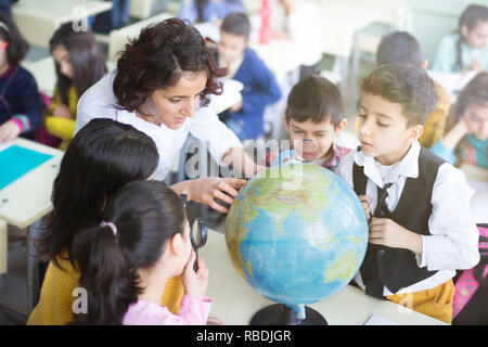 female teacher and students looking the globe with a magnifying glass in the classroom Stock Photo