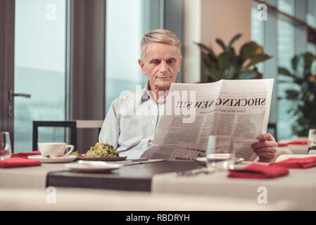 New information. Pleasant old man reading a newspaper while having lunch in a restaurant Stock Photo