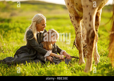 Side view of young mother with little girl in dresses embracing spotty horse on green meadow Stock Photo