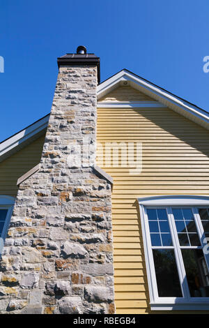 Natural stone chimney on the exterior wall of a yellow cladded with white trim contemporary country style residential home Stock Photo