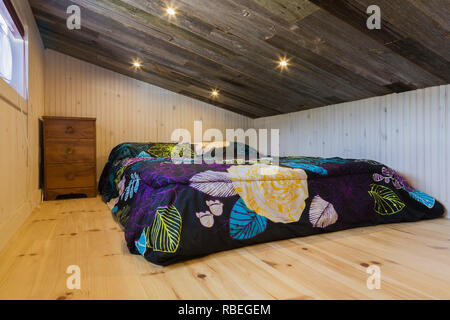 Upstairs bedroom with queen size bed on pinewood floor and old rustic barn wood sloped ceiling inside contemporary 8 x 24 foot mobile mini house Stock Photo