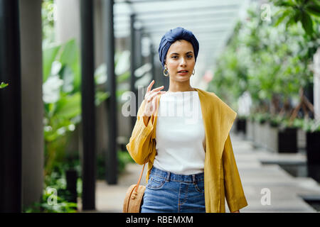Portrait of a tall, beautiful and elegant Middle Eastern Arab woman in a turban and a pastel outfit standing on a street in the city. She is smiling. Stock Photo