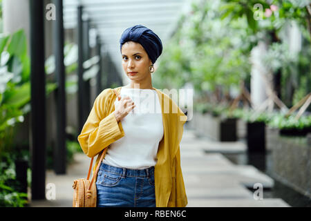Portrait of a tall, beautiful and elegant Middle Eastern Arab woman in a turban and a pastel outfit standing on a street in the city. She is smiling. Stock Photo