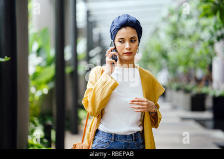 A young and beautiful Middle Eastern woman in a turban hijab frowns as she talks on her smartphone on the street during the day. She looks worried. Stock Photo