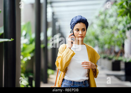 A young and beautiful Middle Eastern woman in a turban hijab frowns as she talks on her smartphone on the street during the day. She looks worried. Stock Photo