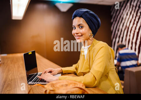 A young Malay Asian woman is typing and working or studying on her laptop computer in a cafe during the day. She is wearing a turban hijab. Stock Photo
