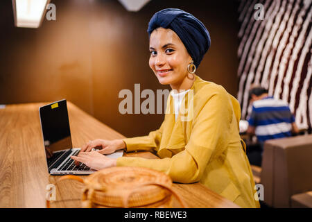 A young Malay Asian woman is typing and working or studying on her laptop computer in a cafe during the day. She is wearing a turban hijab. Stock Photo