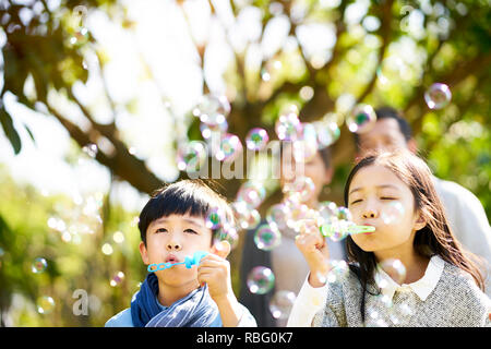 little asian kids boy and girl sister and brother blowing bubbles in a park with parents watching from behind. Stock Photo