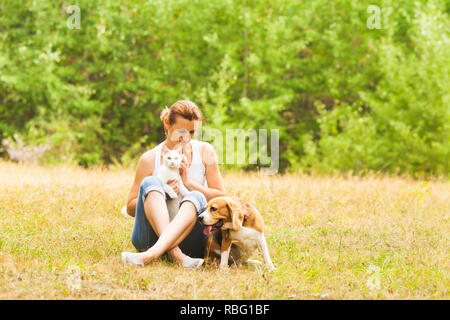 Young nice woman sitting on a ground in the summer meadow holding white fluffy cat on her knees with cute beagle dog standing near, green forest on a  Stock Photo