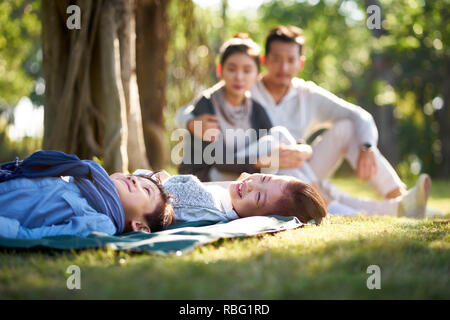 two asian children little boy and girl having fun lying on grass with parents sitting watching in background.