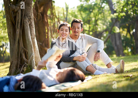 two asian children little boy and girl having fun lying on grass reading a book with parents sitting watching in background.