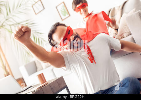 Father and little son wearing superheroe costumes together at home man sitting on floor hand up flying smiling playful close-up while boy sitting on s Stock Photo