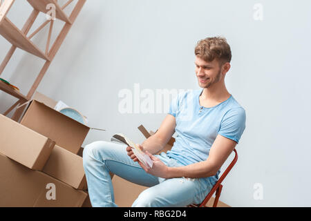 Young blond man moving to new place sitting on ladder looking at palette choosing color laughing cheerful Stock Photo