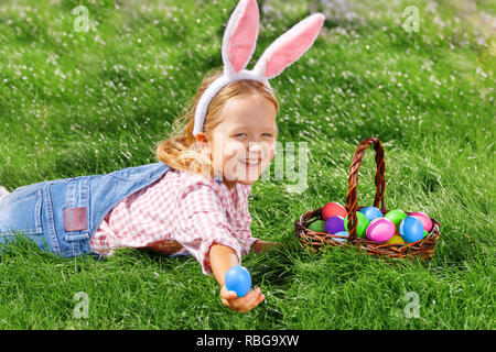 A cheerful little girl preschooler dressed in bunny ears is lying on the lawn with a basket of painted Easter eggs. Happy Easter Stock Photo