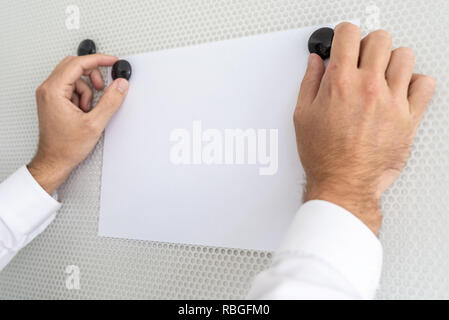 Closeup view of businessman clipping blank white sheet of paper with two black magnets on white magnetic board. Stock Photo
