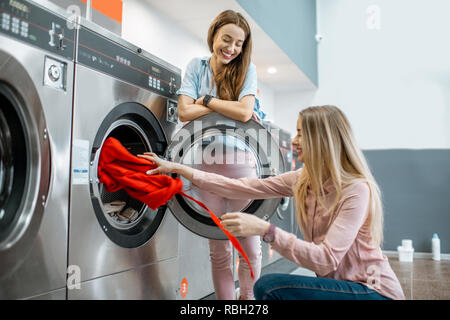 Two cheerful girlfriends having fun loading clothes into the washing machine in the self-service laundry Stock Photo