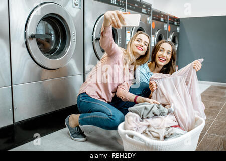 Two cheerful girlfriends making selfie photo with basket full of clothes in the professional laundry Stock Photo