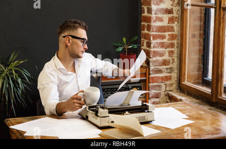 Young writer wearing glasses and white shirt reading new chapter of his book and drinking his morning coffee Stock Photo