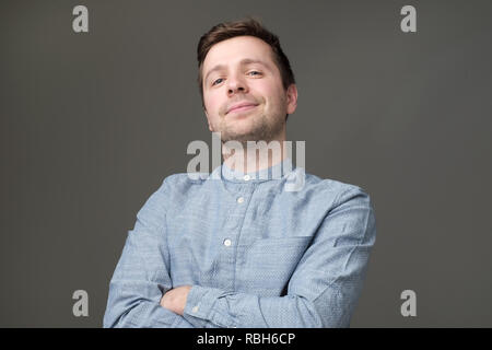 Confident young handsome man in blue shirt keeping arms crossed and smiling while standing against gray background Stock Photo