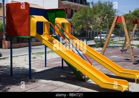 Screened Playground with Two Colorful Slides and Swing. Neighbourhood Public Park with Houses. Unsafe Recreational Activity Area because of the Floori Stock Photo