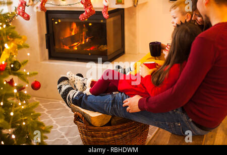 Family sitting on the branch and warm their foot near the fireplace. Chrismtas time, family time together with mugs of hot cocoa or tea Stock Photo