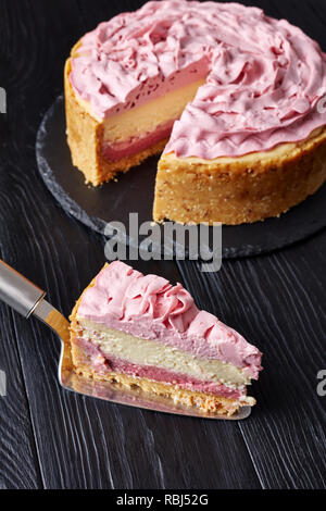 piece of a white chocolate raspberry cheesecake on a cake shovel and sliced cheesecake at the background on a black stone tray on wooden table, vertic Stock Photo