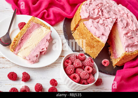 portion of a white chocolate raspberry cheesecake on a plate and sliced cheesecake on a black stone tray with cloth and frozen raspberries on a white  Stock Photo