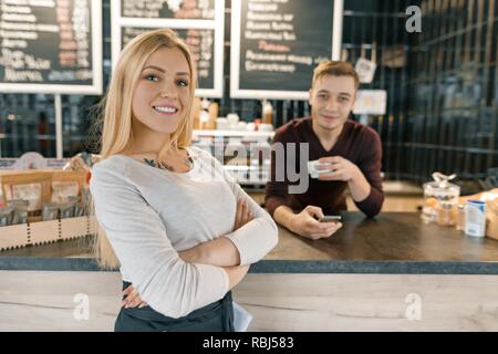 Young smiling couple of coffee shop workers, man and woman posing near the bar counter and coffee machine with cup of fresh coffee. Teamwork, staff, s Stock Photo