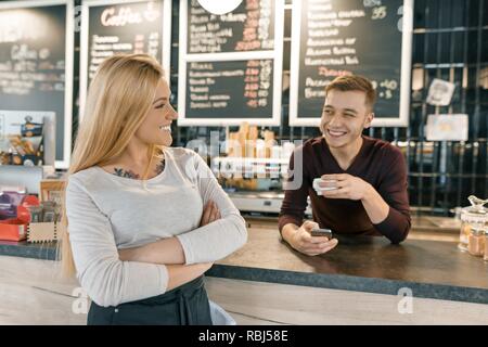 Young smiling couple of coffee shop workers, man and woman posing near the bar counter and coffee machine with cup of fresh coffee. Teamwork, staff, s Stock Photo