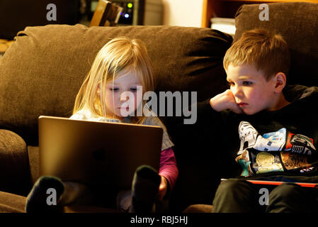 A little girl (4 yrs old) sat on a sofa using a laptop computer while her brother (6 yrs old) watches.