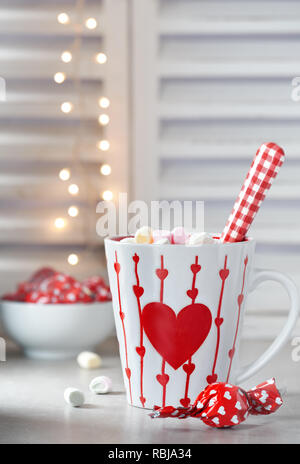Hot chocolate with marshmallows, red heart on the cup, winter background with lights out of focus. Winter or Valentine's day background. Stock Photo