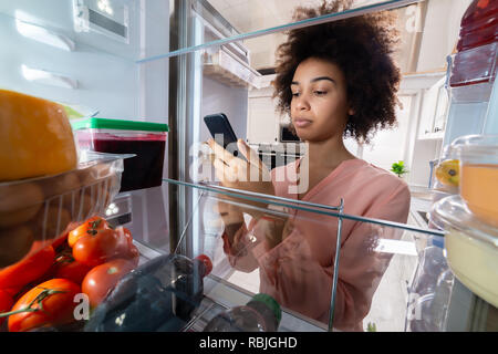 Happy Young Woman Standing In Front Of Refrigerator With Food Items Using Mobile Phone Stock Photo