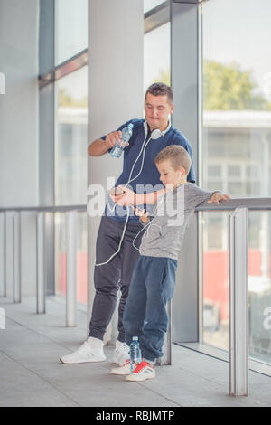 Little boy and his father taking a break, drinking water and listening to the music together in a sports center. Father and son spend time together an Stock Photo